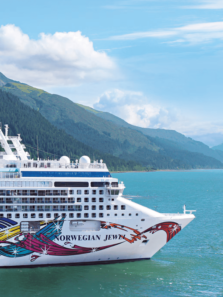 Luxurious cruise ship Norwegian Jewel sailing through scenic fjords with mountains in the background.