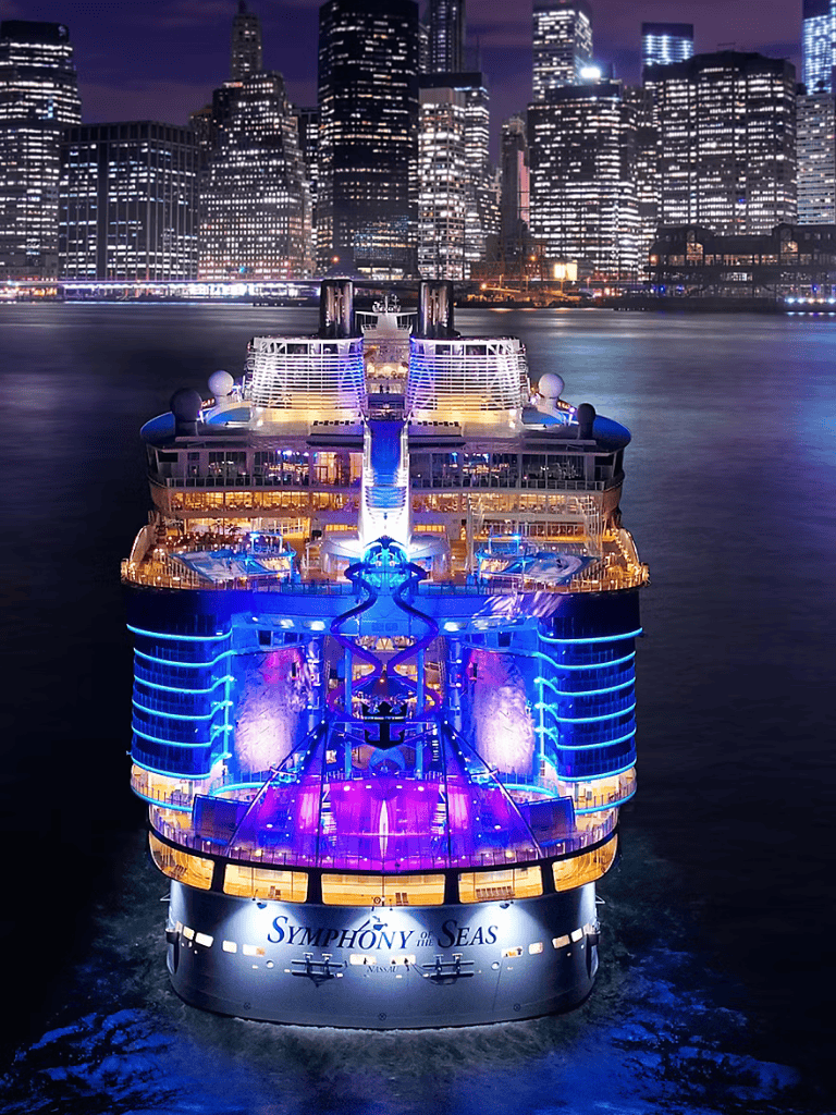 Luxurious Symphony of the Seas cruise ship sailing through a vibrant city skyline at night.