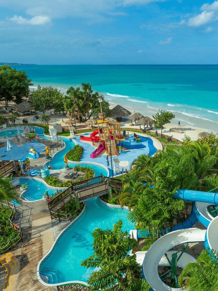 Vibrant water park overlooking a tropical beach with slides, lazy river, and lush greenery.