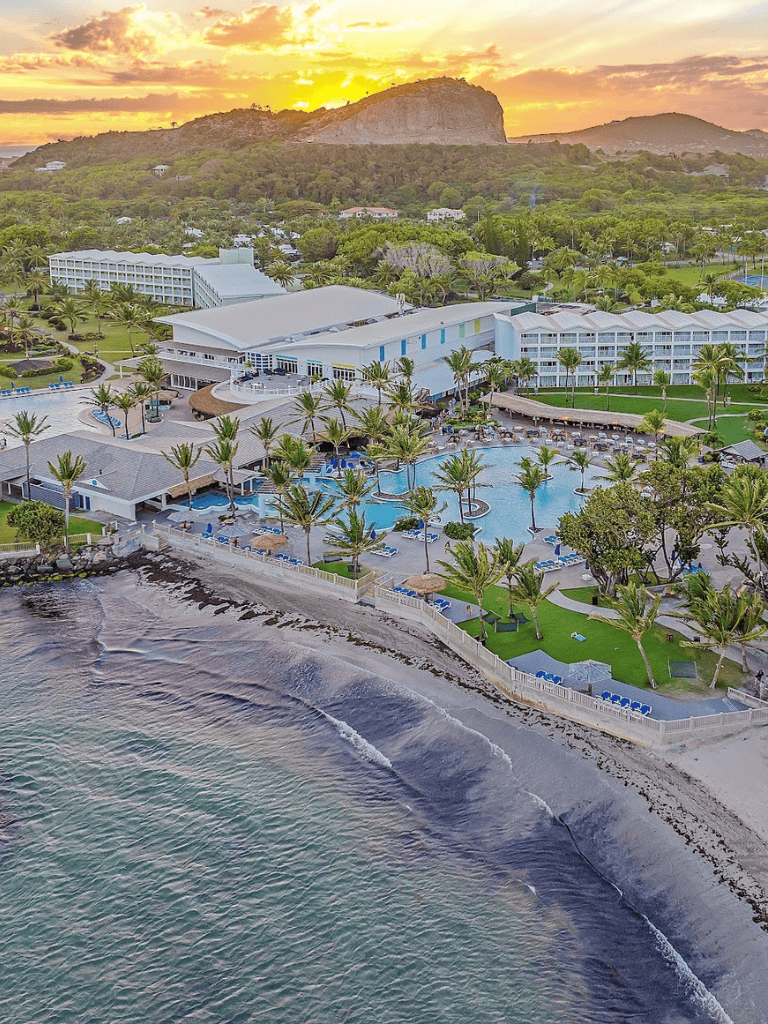 Oceanfront resort with pool, palm trees, and mountain view at sunset.