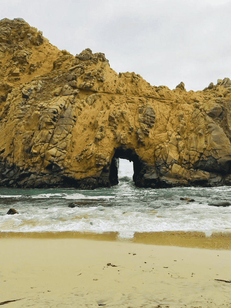 Seaside rock formation with natural arch on sandy beach, coastal scenery, ocean waves, cloudy sky, California coastline.
