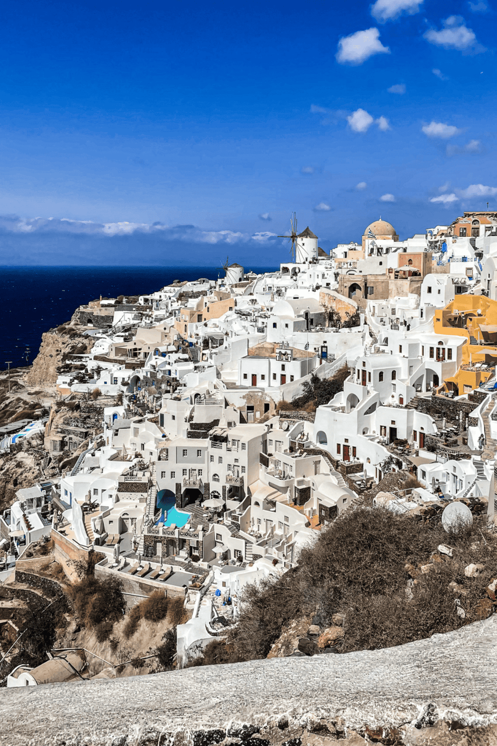 White-washed cliffside village in Santorini with iconic windmills and blue ocean backdrop.