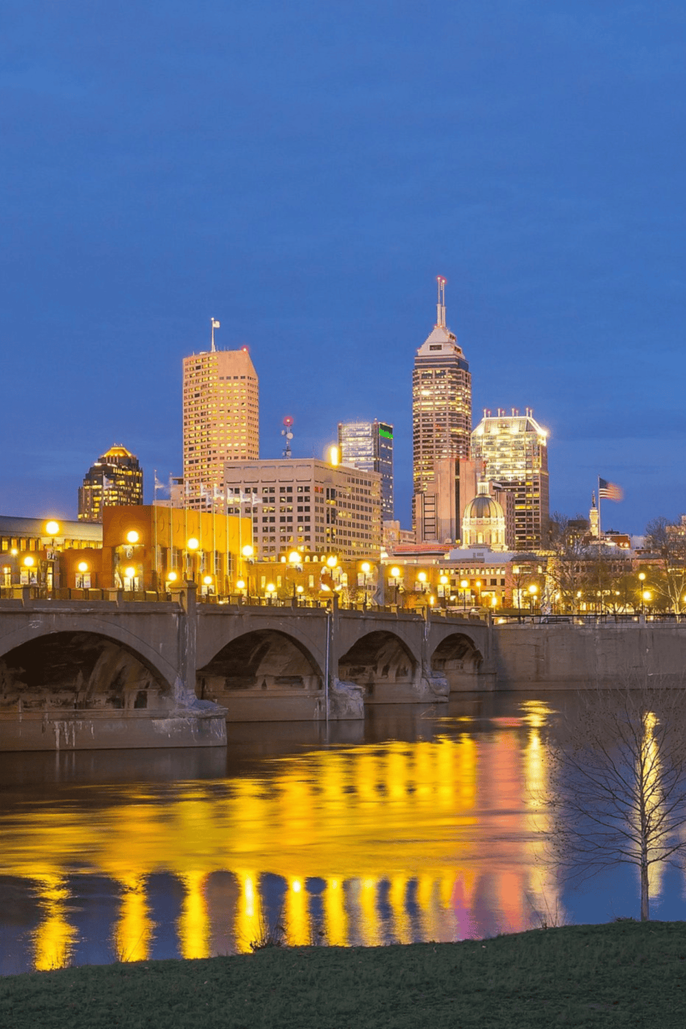 Downtown Indianapolis skyline at dusk with city lights reflecting on the river.