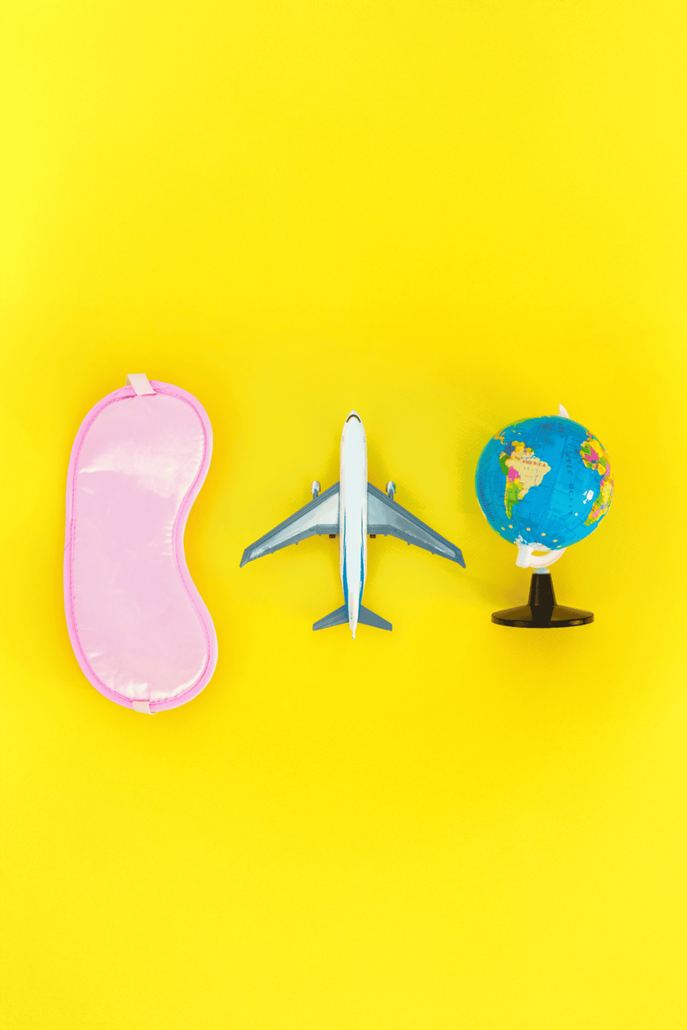 Bright travel accessories on yellow background, including a pink sleep mask, airplane model, and globe, for adventure planning and directions.
