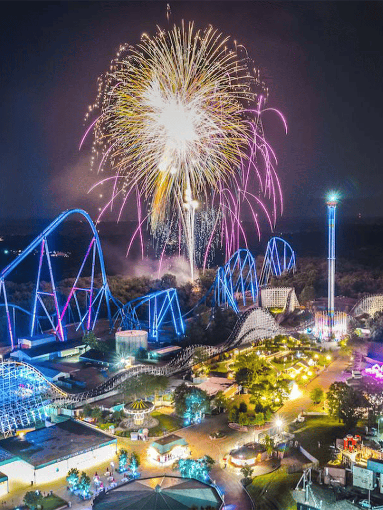 Colorful fireworks over a vibrant amusement park at night, with roller coasters and rides illuminated.