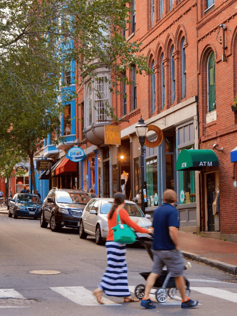 Colorful downtown street with shops, pedestrians, and parked cars, featuring historic brick buildings and a lush tree.