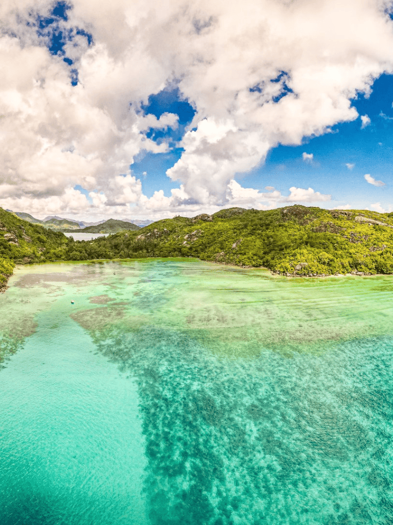 Vibrant turquoise lake surrounded by lush green mountains and blue sky with clouds.