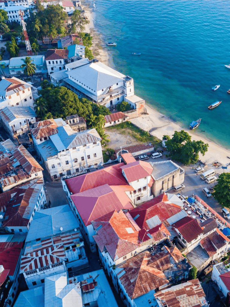 Colorful coastal town with beach and boats, vibrant rooftops, lush greenery, and serene waters.