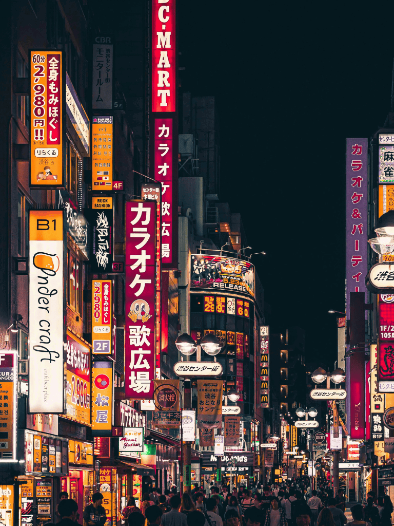 Bright Tokyo city street at night with illuminated signs and crowded pedestrians, neon-lit shops and entertainment venues.