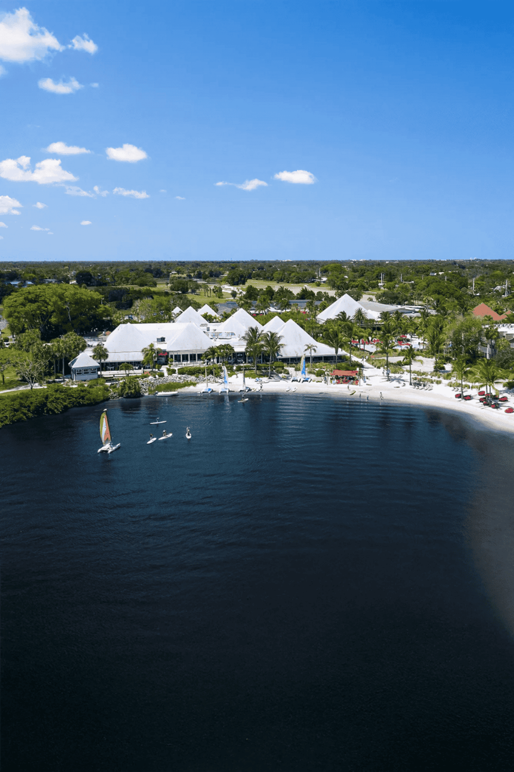 Scenic waterfront with sailboats and tropical resort, vibrant blue sky, lush greenery, and white tents for events or dining.