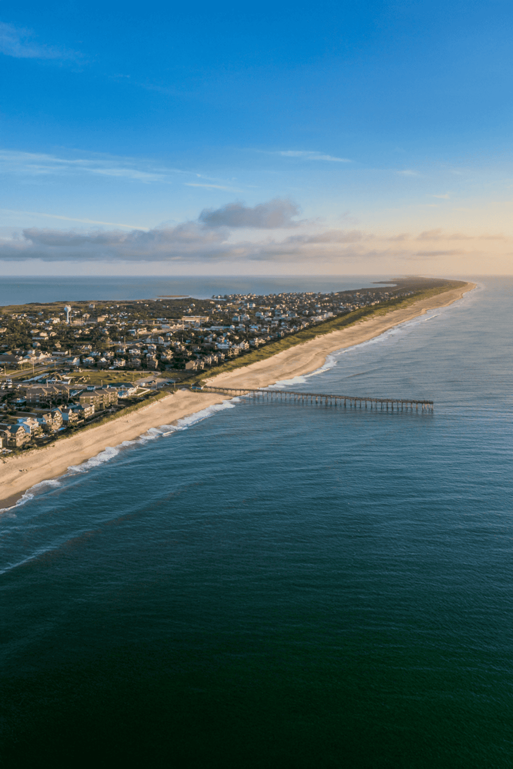 Aerial view of coastal town with pier, sandy beach, and ocean waves at sunset, showcasing scenic seaside destination.