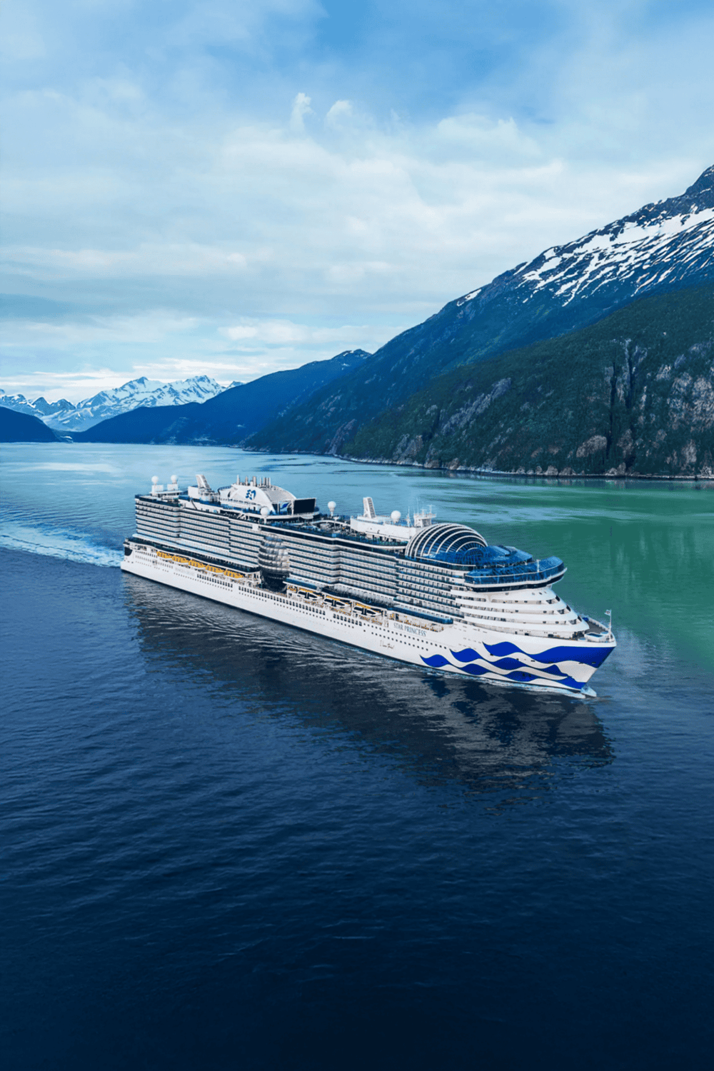 Cruise ship sailing through scenic fjords with snow-capped mountains in the background.