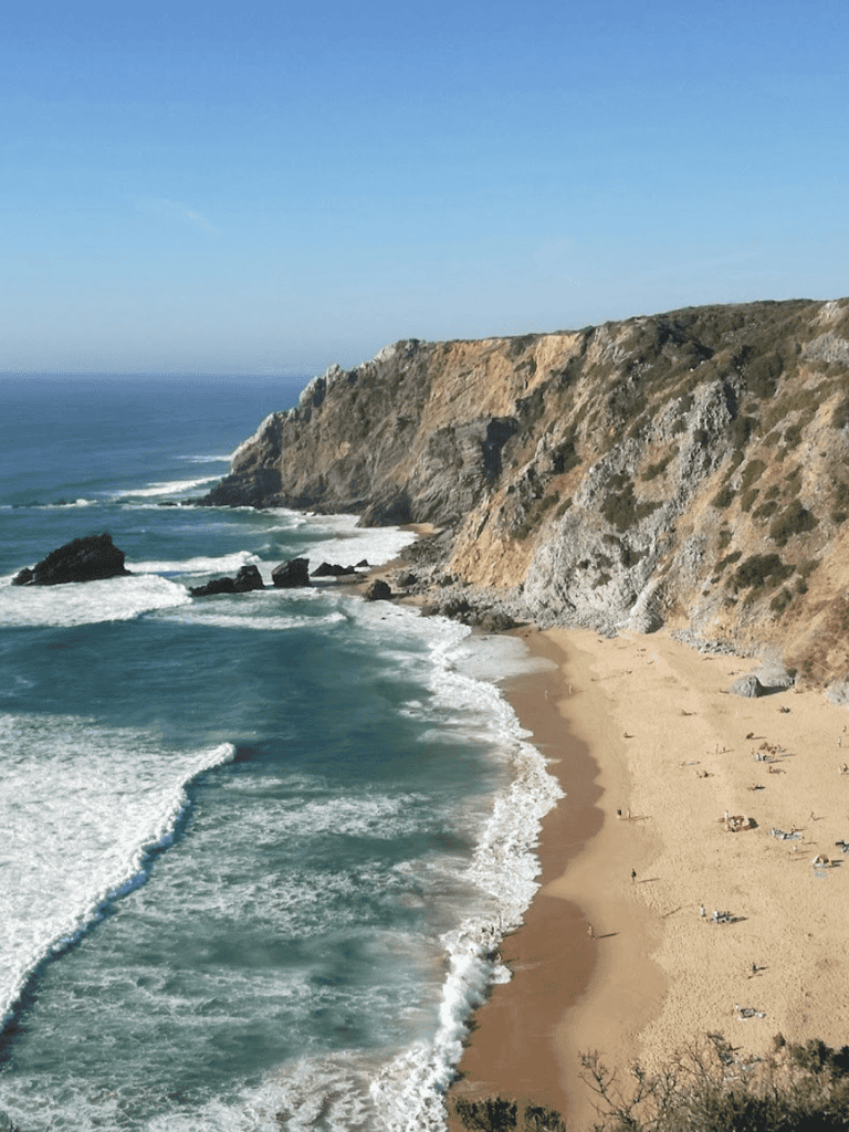 Gentle waves crashing on sandy beach with rugged cliffs under clear blue sky.