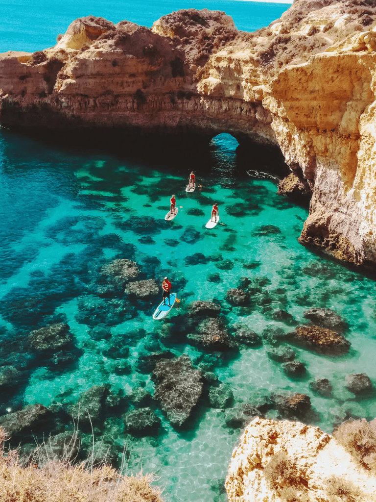 Colorful cliffside coastal scenery with paddleboarders exploring turquoise waters and natural rock formations.