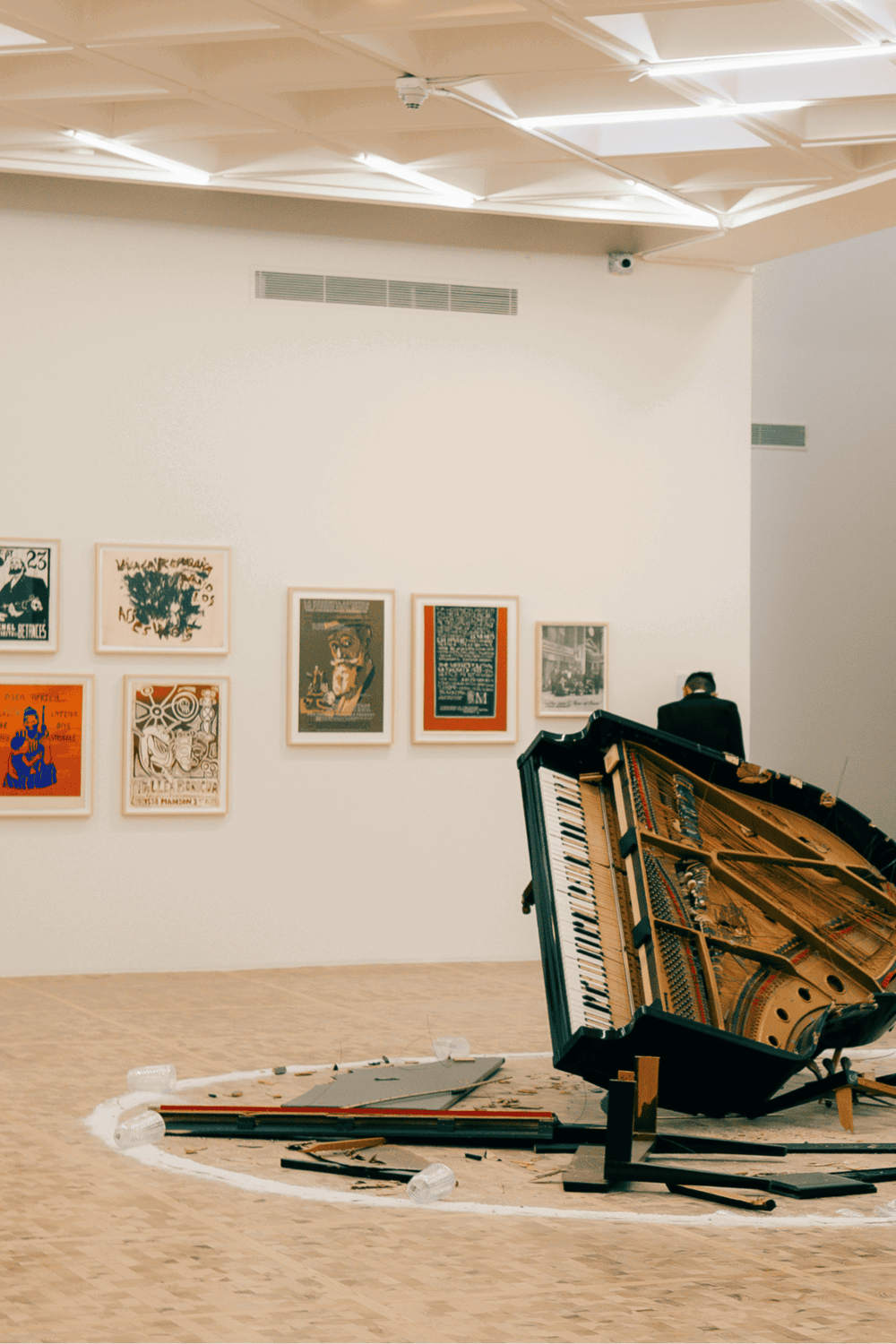 Broken grand piano in art gallery with scattered debris, modern artwork on walls, and a person observing.