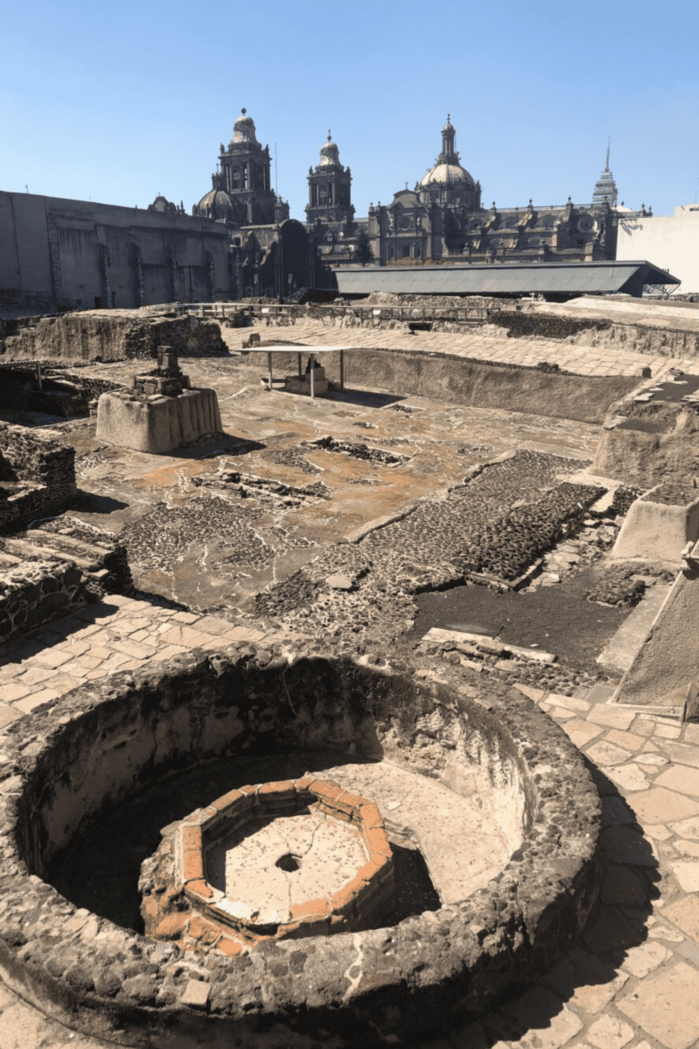Ancient Mayan ruins with historic stone structures and a colonial cityscape in the background.