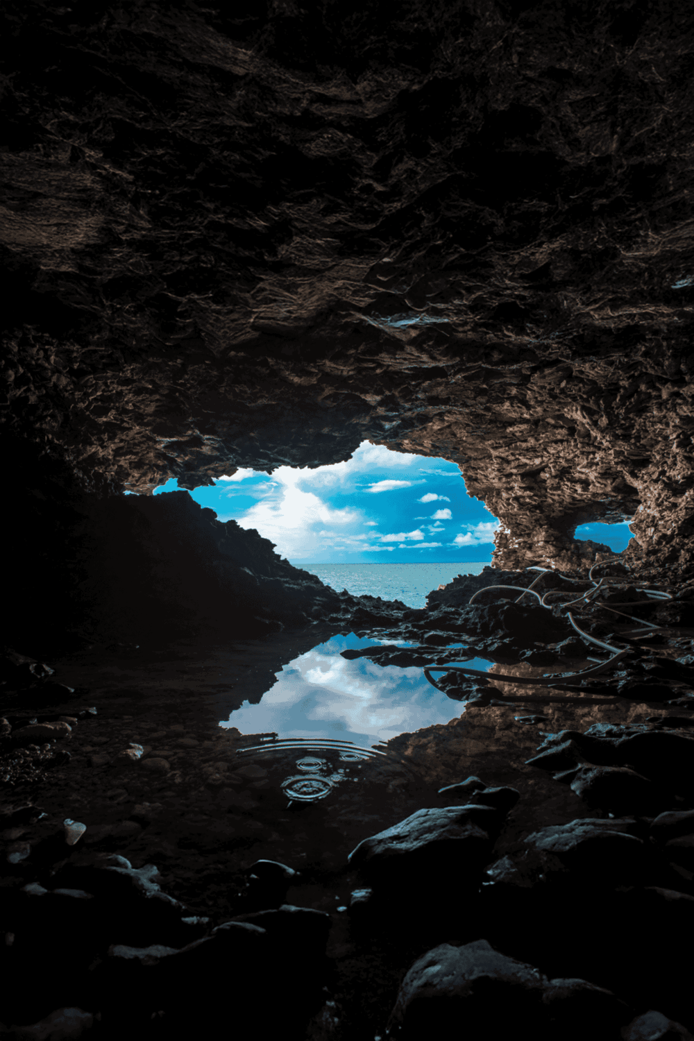 Bright view of the ocean through a rocky cave opening, reflecting blue sky and clouds on water surface.