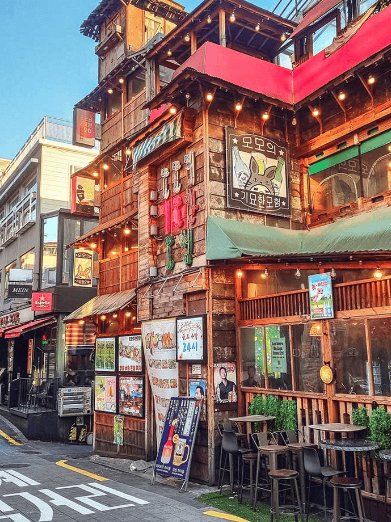 Vintage-themed bar on bustling city street with outdoor seating and neon signs.