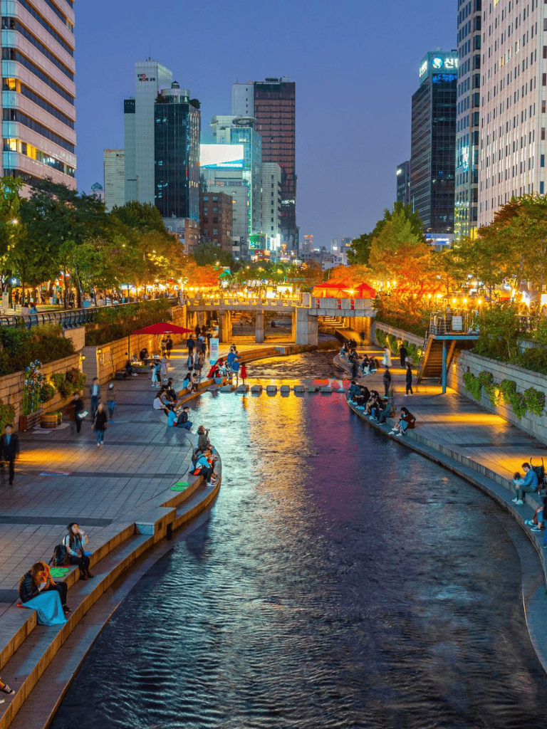 Busy city park at dusk with people sitting by water, surrounded by illuminated skyscrapers in Seoul, South Korea.