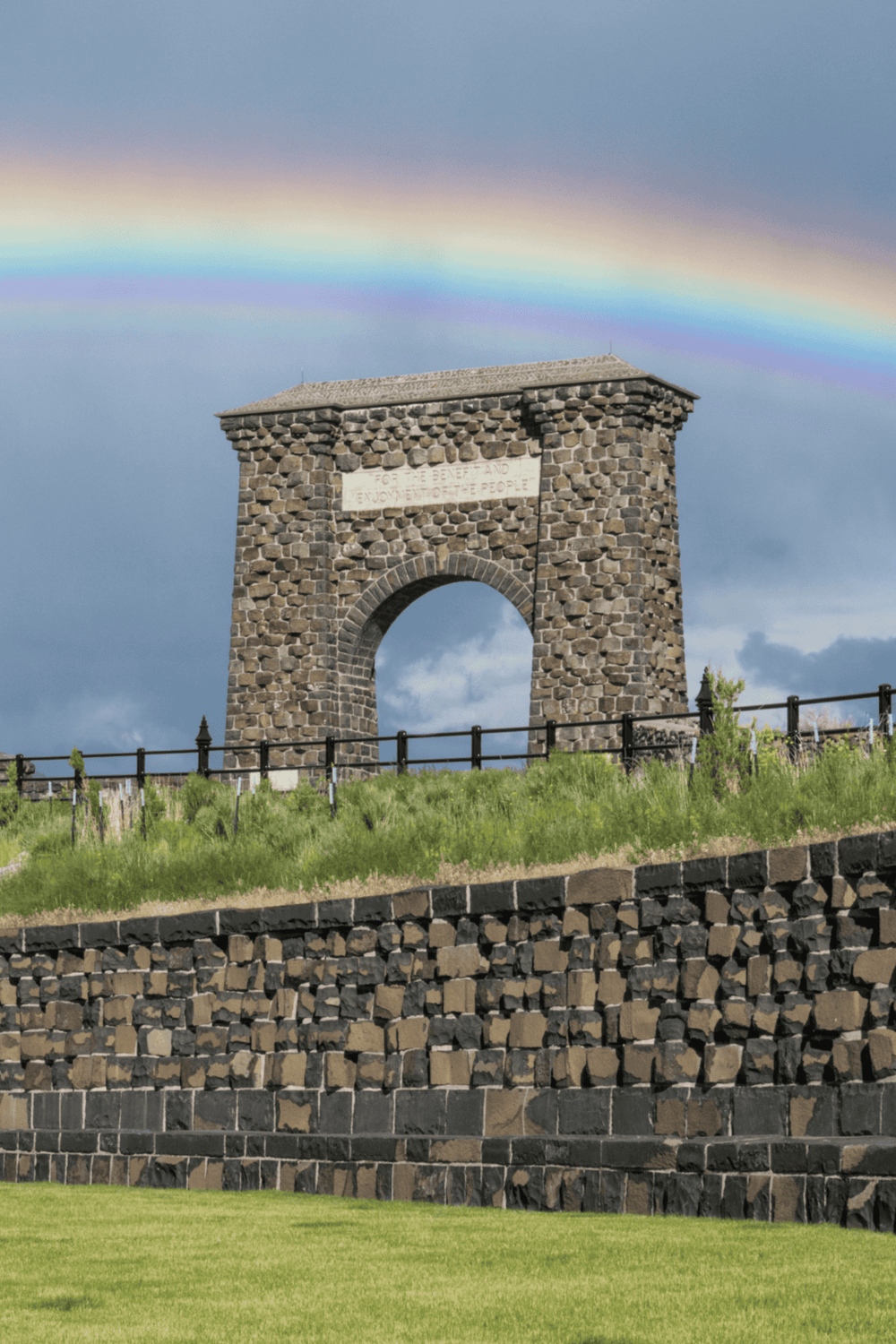 A rainbow arches over a historic stone arch monument on a grassy hill.