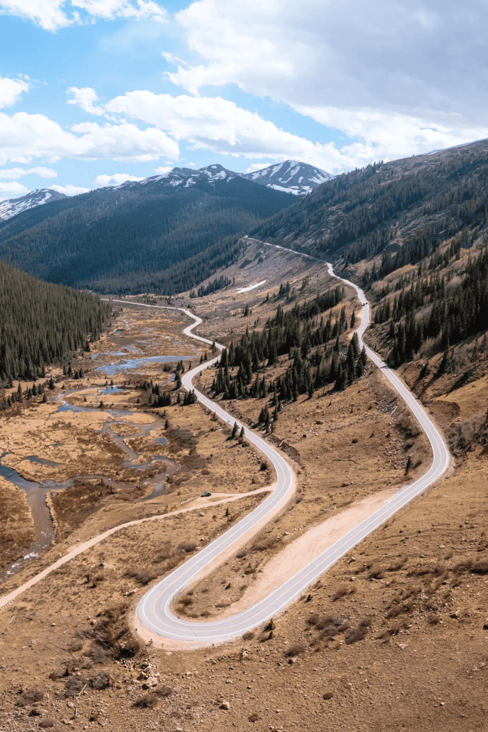 Winding mountain road through scenic valley with snow-capped peaks in the distance.