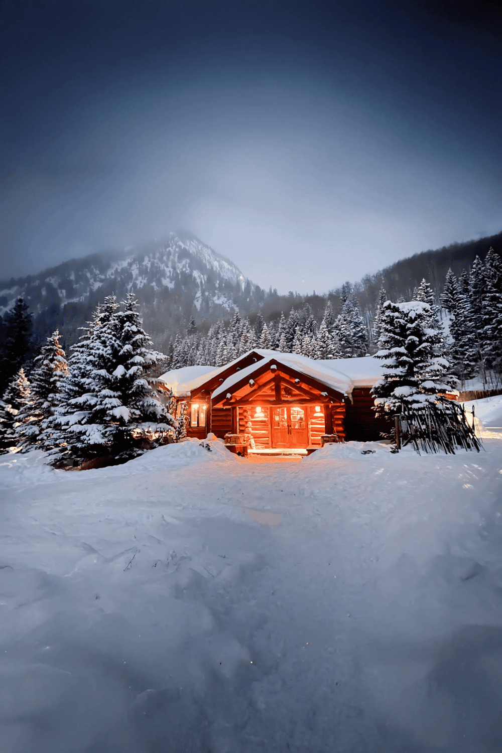 Snow-covered mountain cabin at dusk with warm interior lights, surrounded by pine trees, winter landscape, mountain scenery, cozy retreat, QuestForDirections.