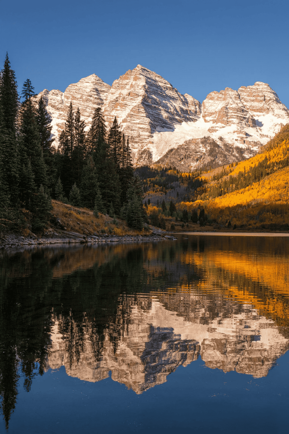 Majestic snow-capped mountains with autumn-colored trees reflected in calm water, scenic outdoor landscape.