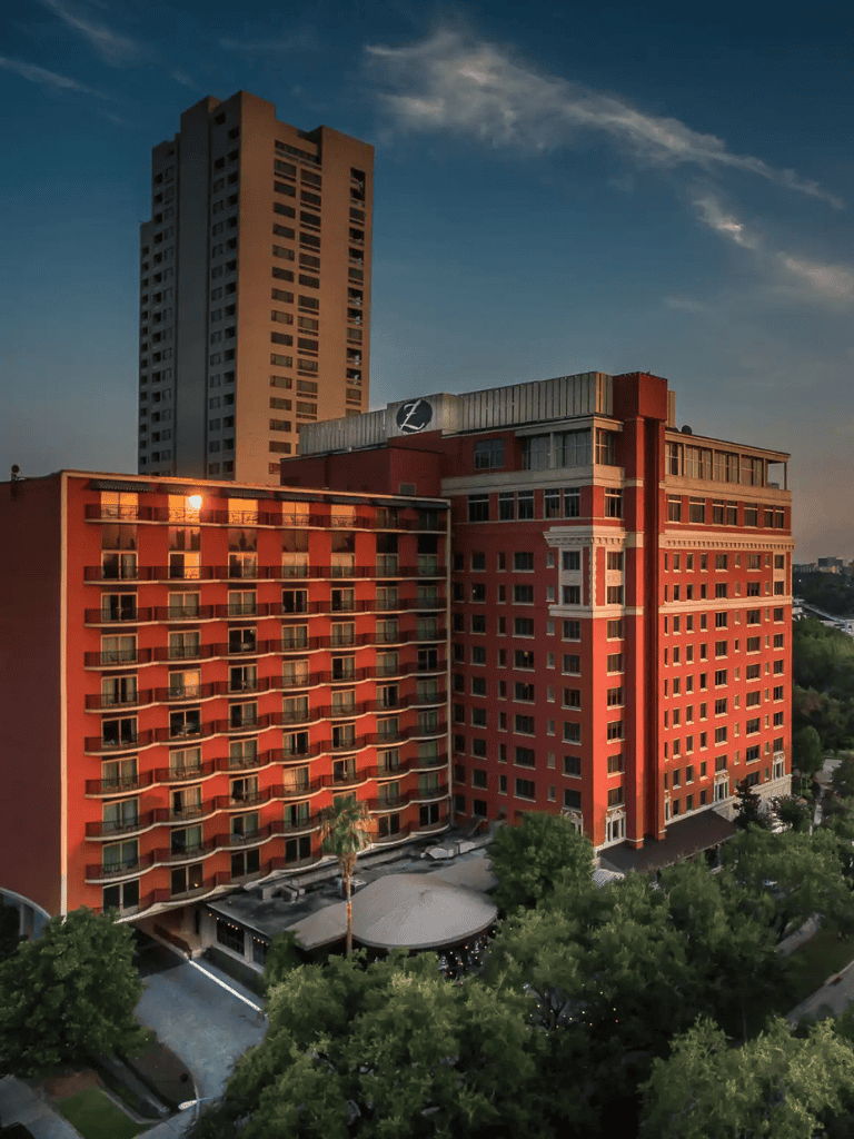 Modern hotel building with red exterior and cityscape backdrop at sunset.