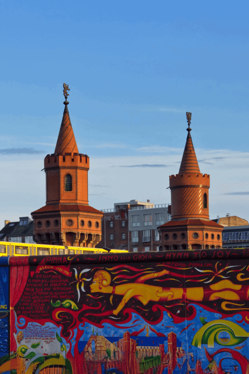 Colorful street art mural with two red brick towers and modern buildings in the background.
