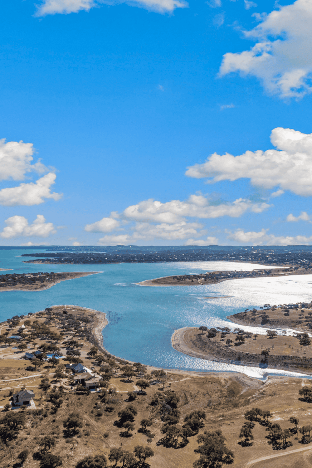 Aerial view of a serene lake with flowing water, surrounded by dry land and scattered trees under a bright blue sky.