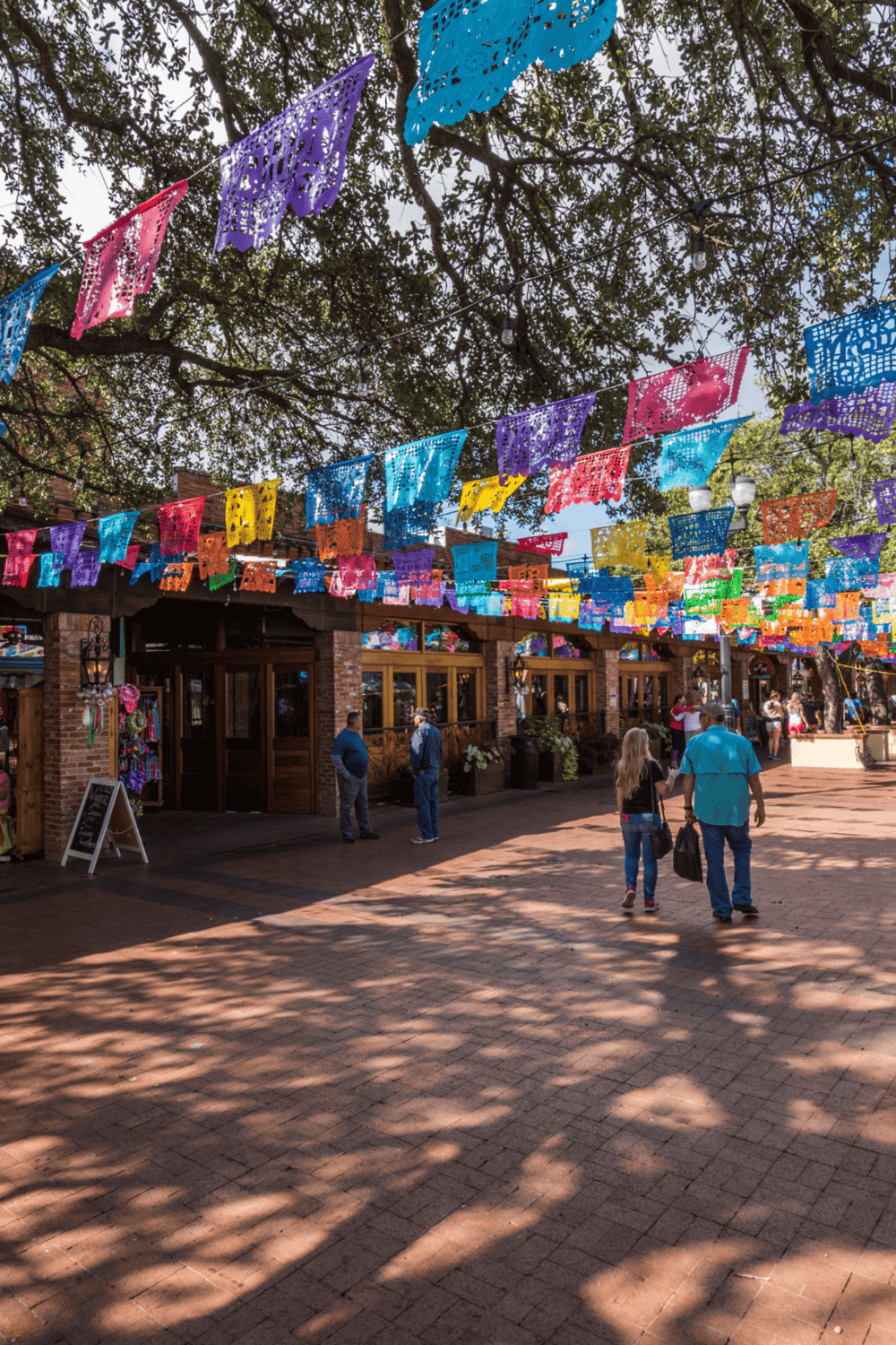 Colorful papel picado banners hanging above a lively outdoor marketplace with people walking and shopping.