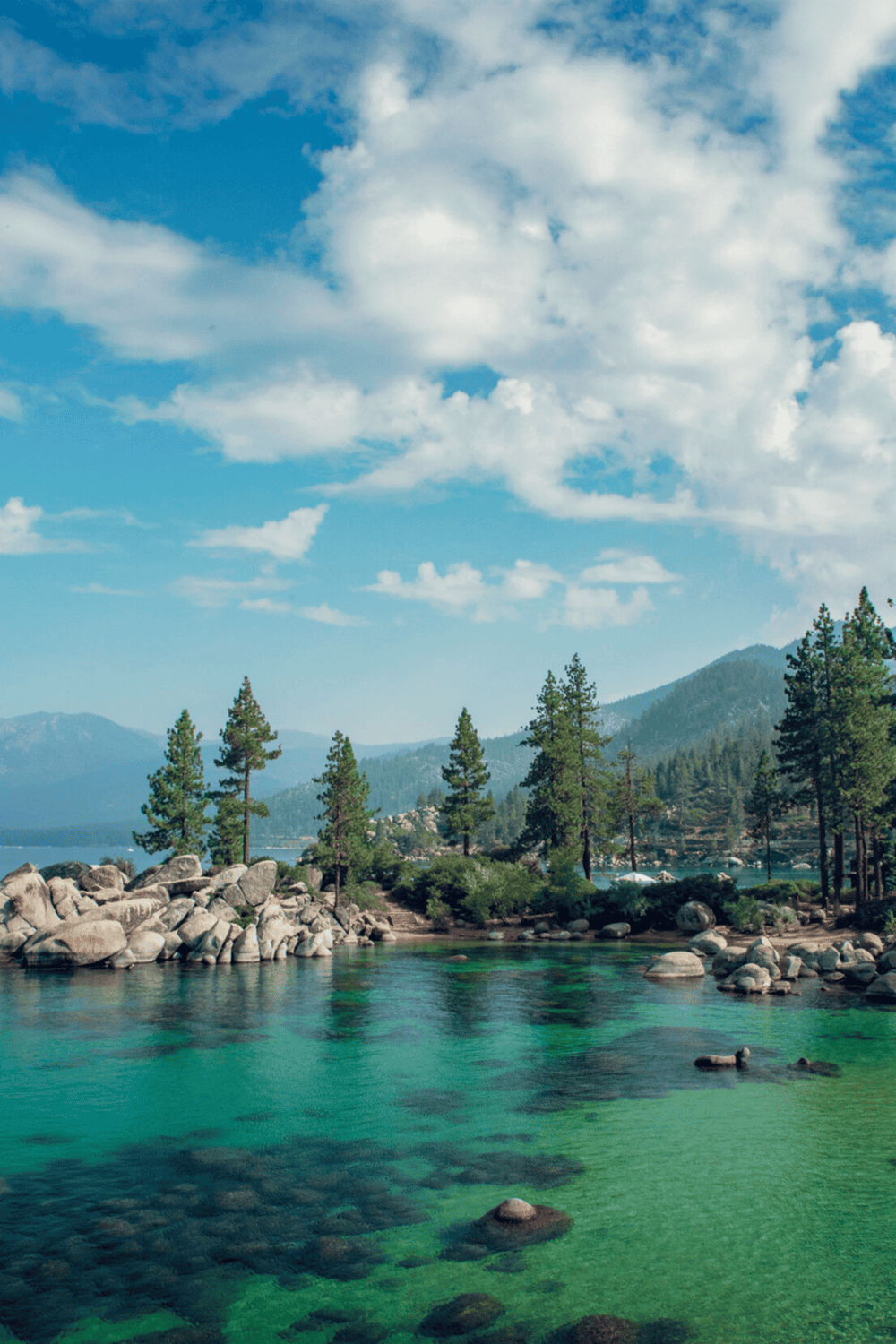 Tranquil lake surrounded by pine trees and mountains under a blue sky with clouds.