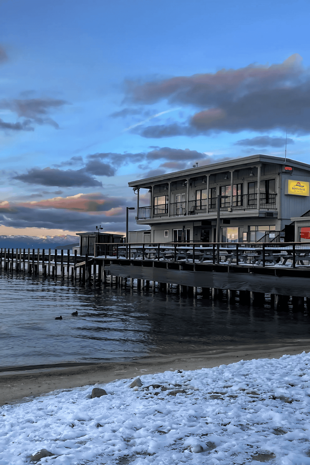 Waterside restaurant on pier with mountain view, sunset sky, and snow at the shoreline.