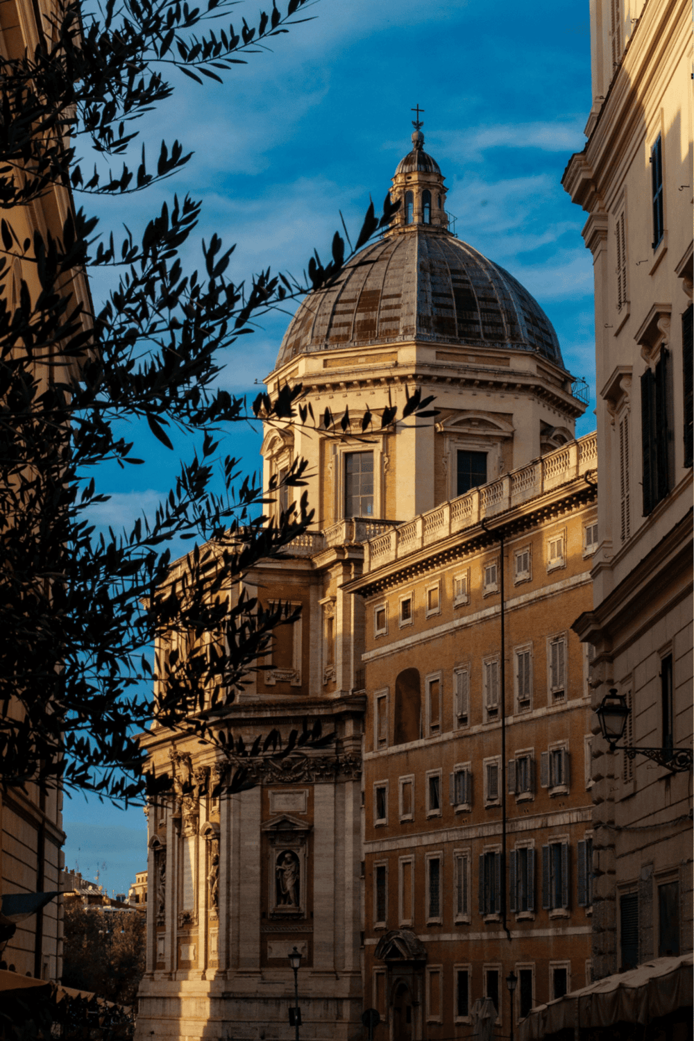 Ancient Roman church with a large dome, basking in golden sunlight, framed by historic buildings and dark foliage.