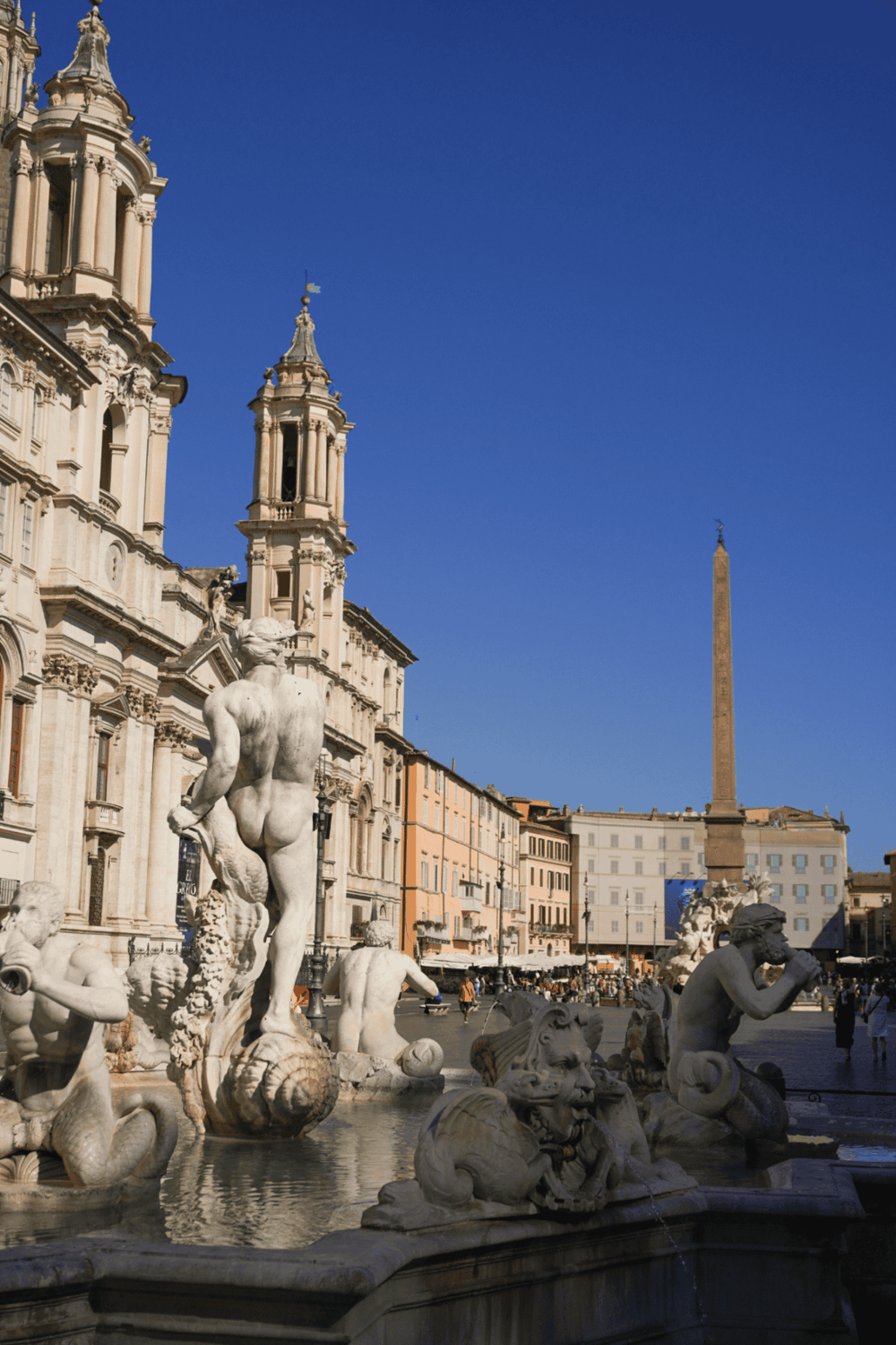 Intricate Baroque fountain in Piazza Navona, Rome, with historic buildings and obelisk in the background.