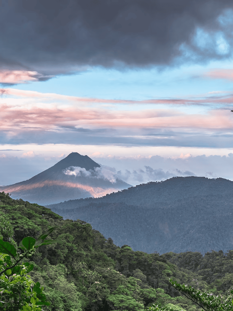 Lush green rainforest with volcano under dramatic cloudy sky at sunrise or sunset.