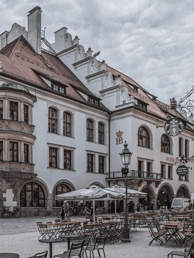Bavarian-style building with outdoor seating and Starbucks sign, cobblestone plaza, cloudy sky.