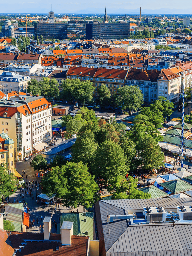 Aerial view of a vibrant European city square with colorful buildings, lush trees, and bustling outdoor markets.