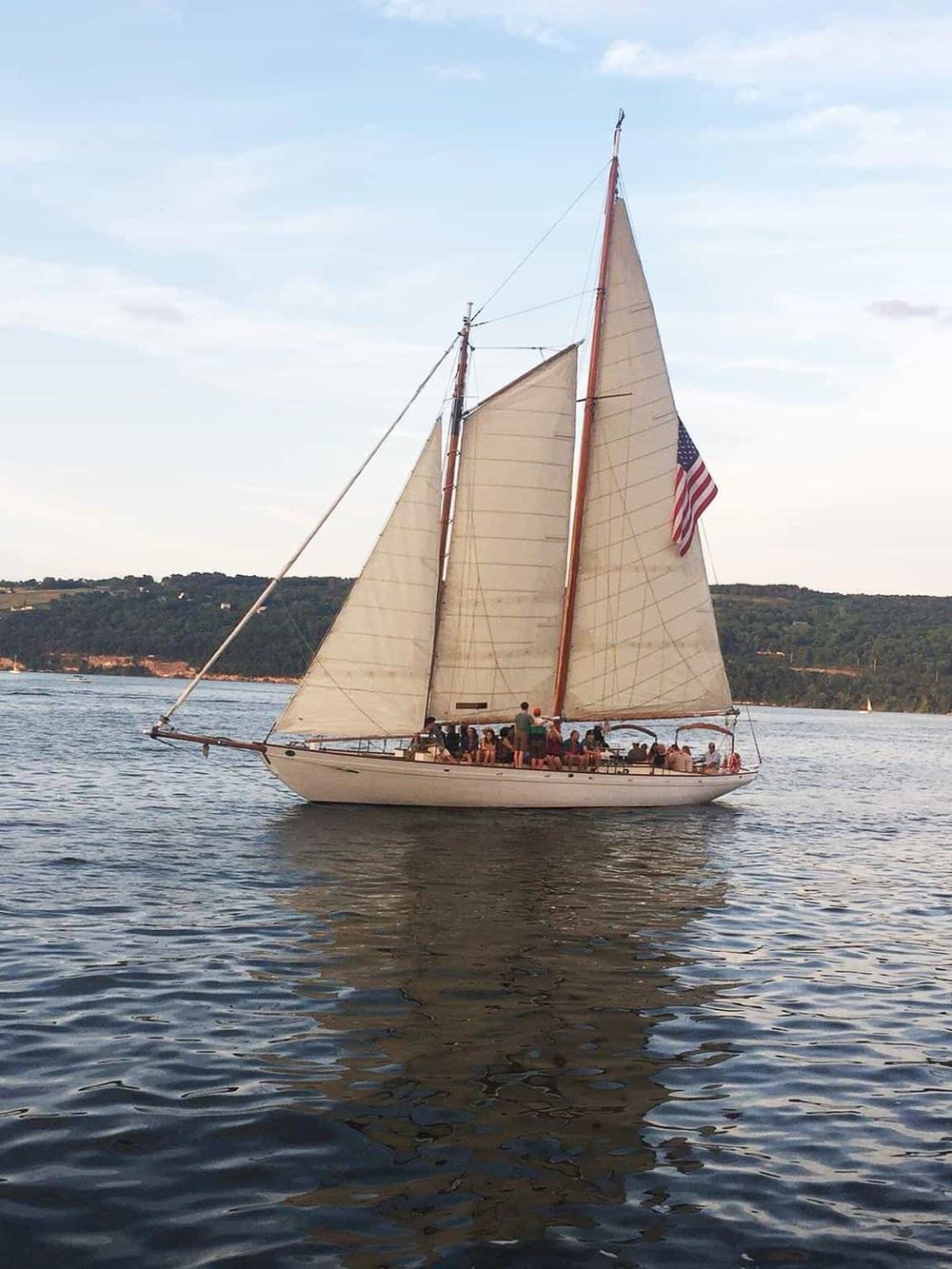 Sailing boat on water with sails and American flag, scenic harbor view with distant hills.