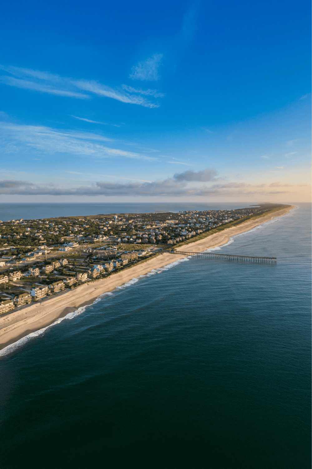 Aerial view of a coastal town with a sandy beach, ocean pier, and residential area under blue sky.