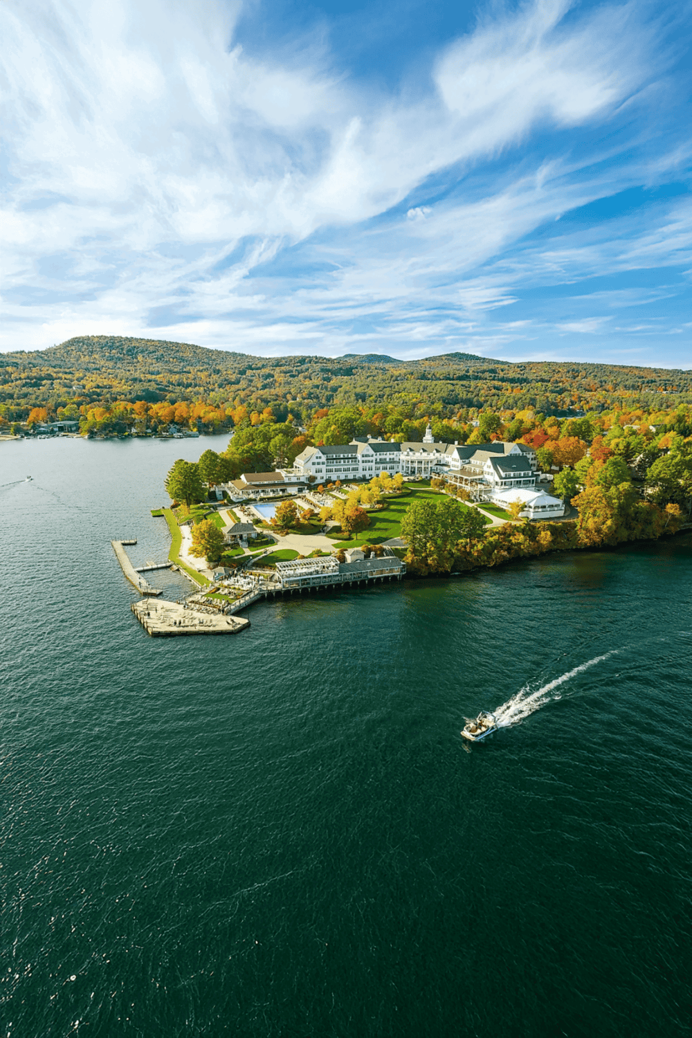 Aerial view of a lakeside resort surrounded by colorful autumn foliage and mountains, with boat on water.
