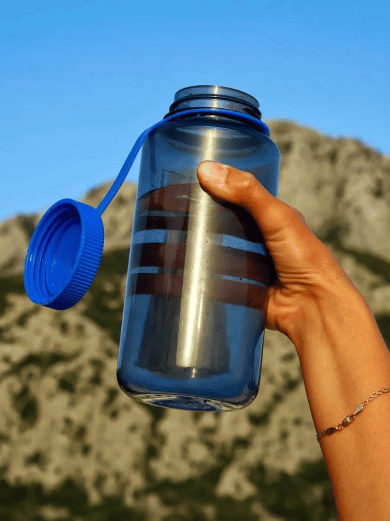 Hand holding a reusable water bottle with an outdoor mountain background.
