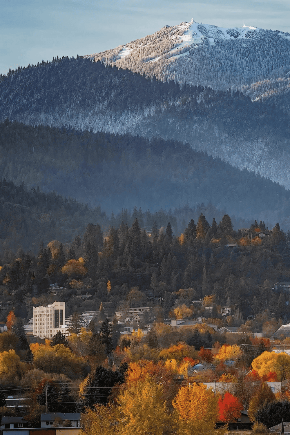 Vast mountain landscape with a town at the foothills, showcasing stunning autumn foliage and snow-capped peaks in the distance.