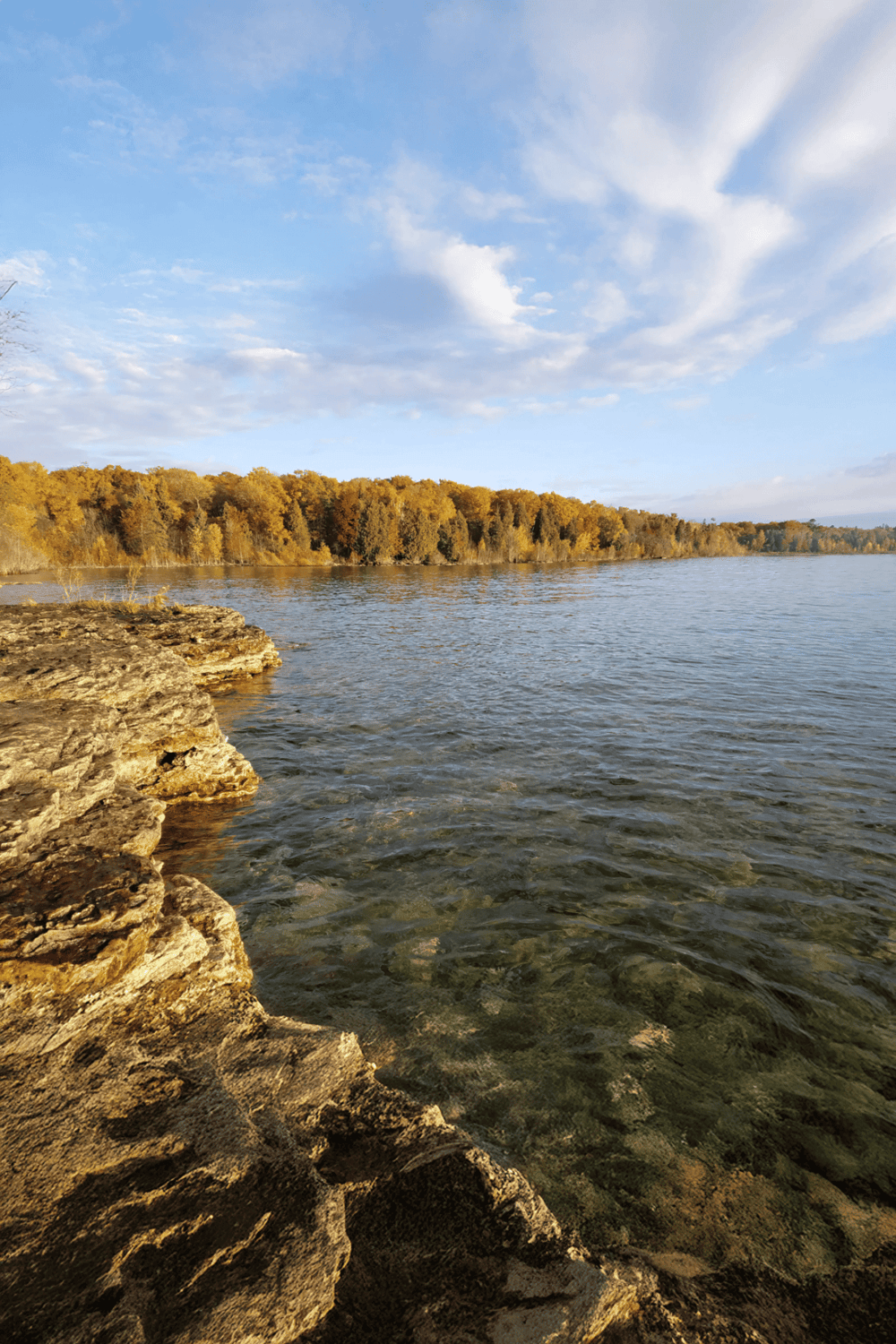Calm river shoreline with rocky bank and autumn trees under a blue sky, scenic outdoor nature view.