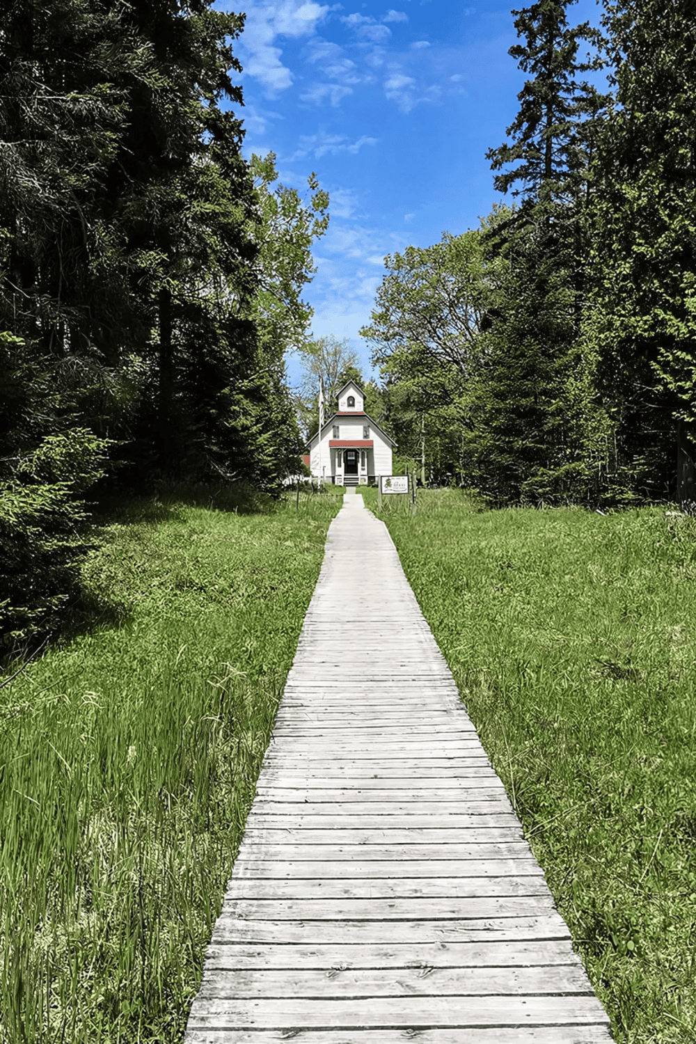A white church surrounded by trees along a wooden pathway on a bright, sunny day.