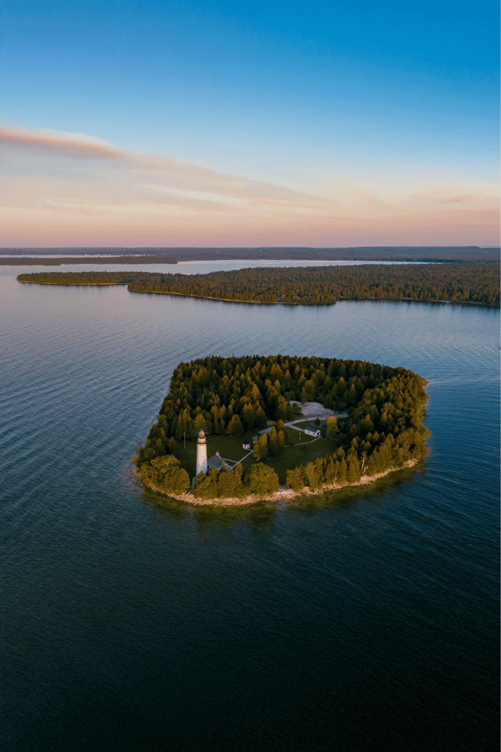 Secluded island with lighthouse and dense trees surrounded by calm water at sunset.