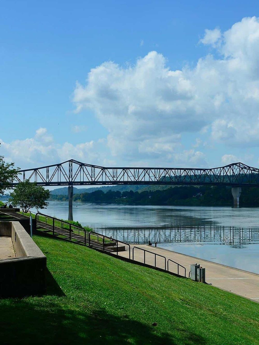 Scenic view of a bridge over a calm river with a park and pathway in the foreground, under a partly cloudy sky.