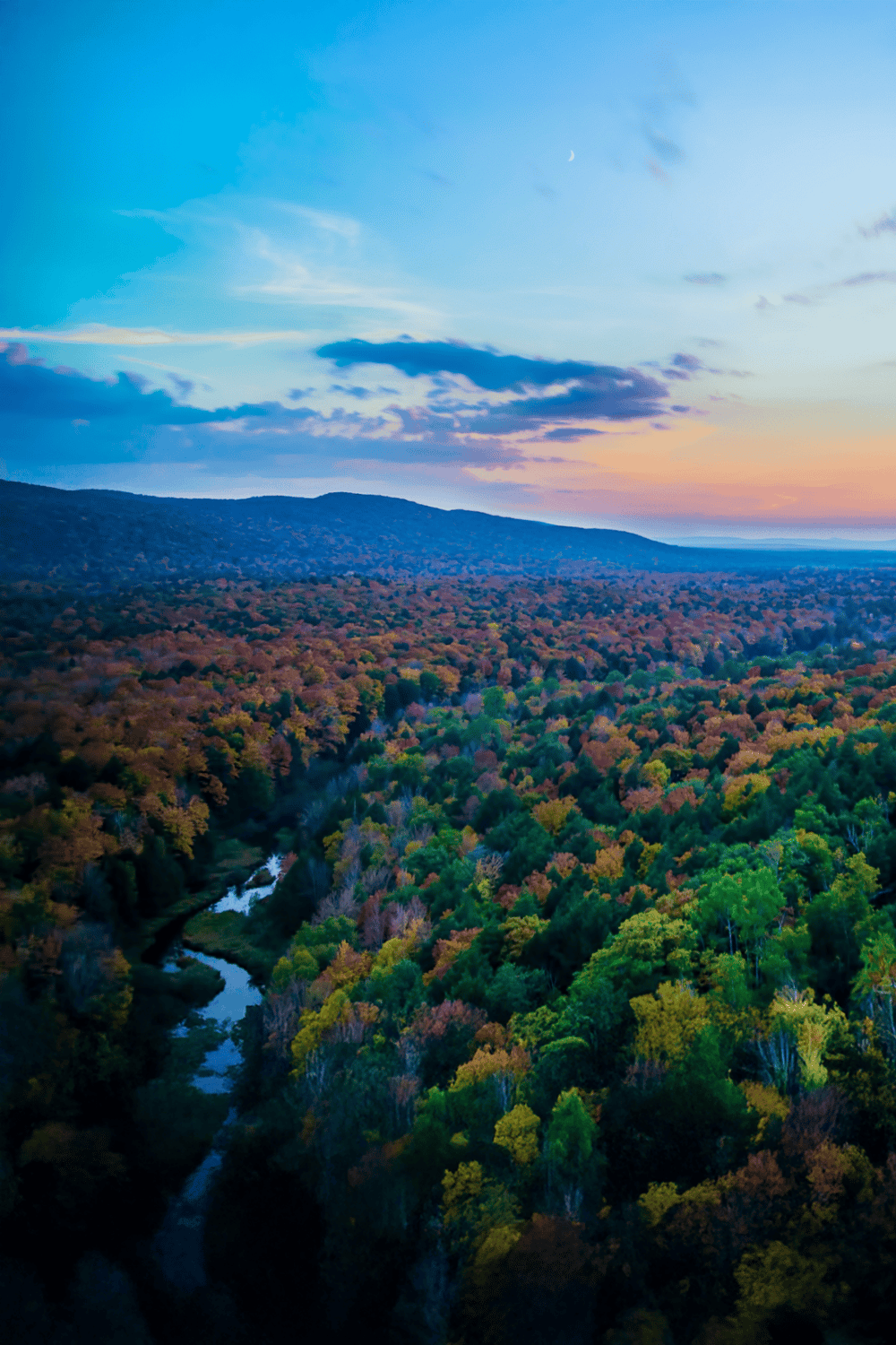 Rolling hills and vibrant autumn forest with a winding stream under colorful sky at dusk.