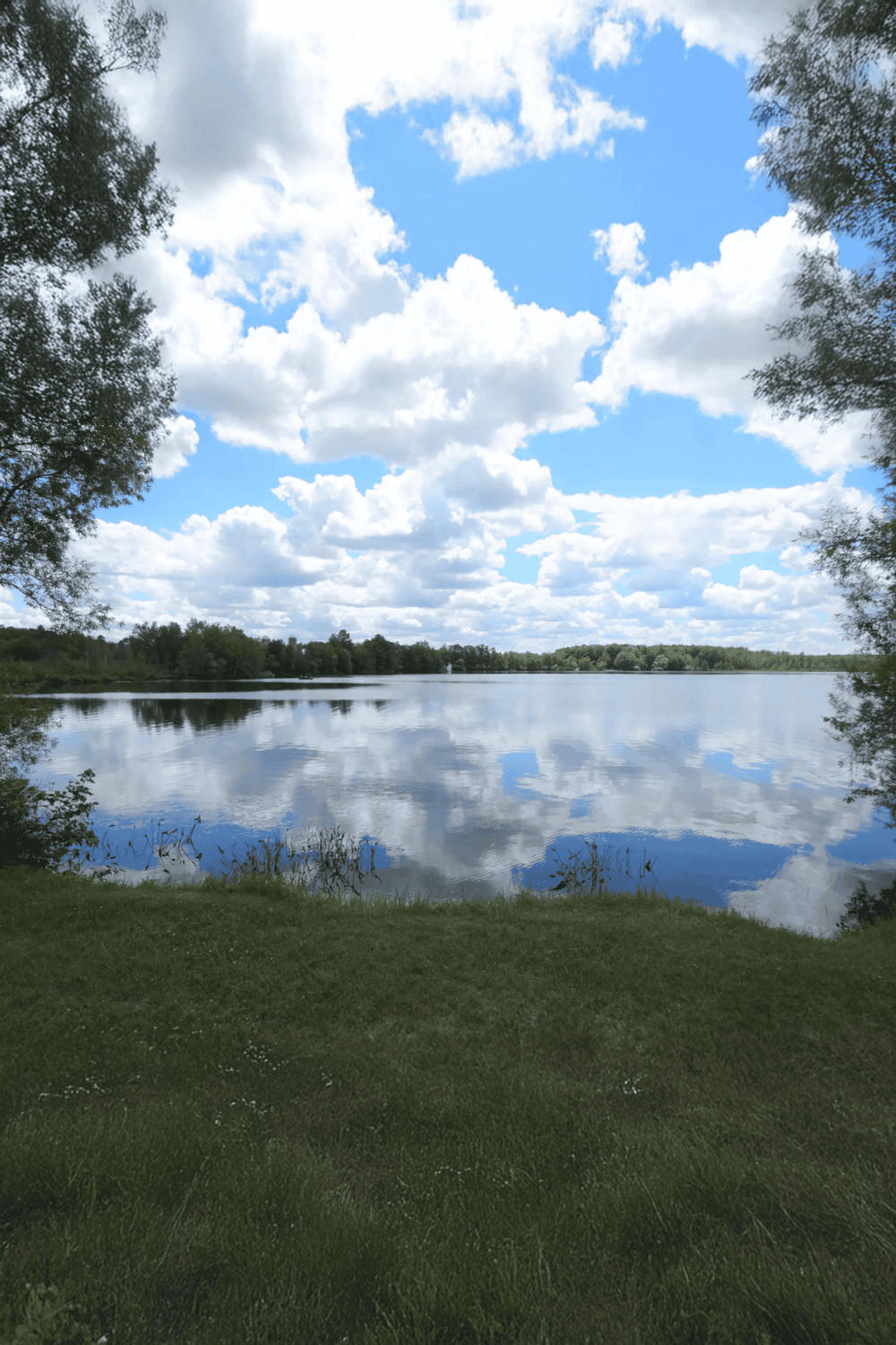 Serene lake with reflections of blue sky and clouds, surrounded by trees and lush greenery.