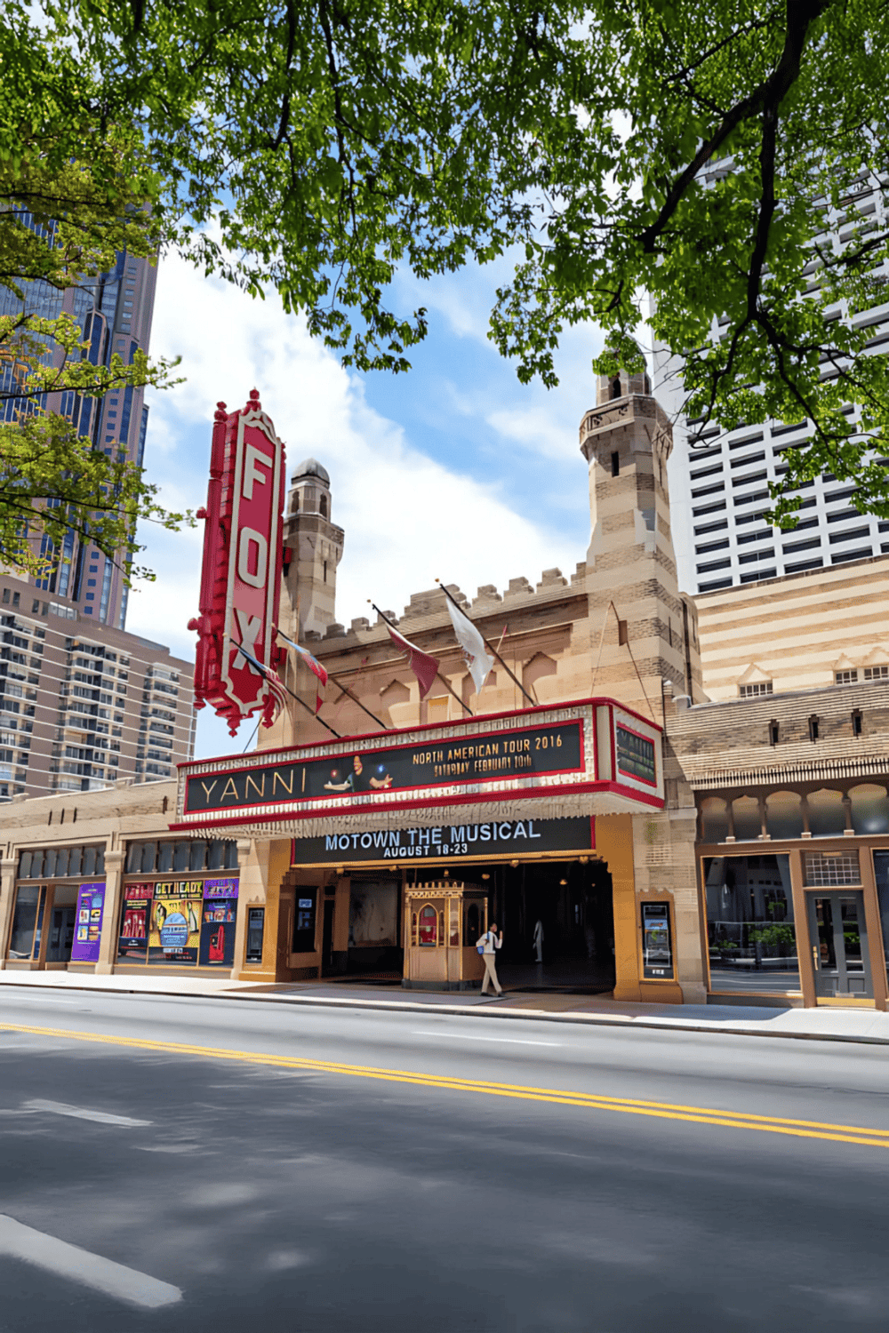 Historic theater building with marquee advertising musical, surrounded by modern skyscrapers and lush trees.